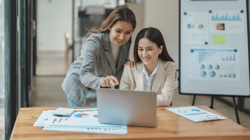 Duas mulheres estão trabalhando em um computador, estando atrás um cavalete como suporte de relatórios gerenciais com gráficos, fornecidos rapidamente pelo BPO Financeiro. 